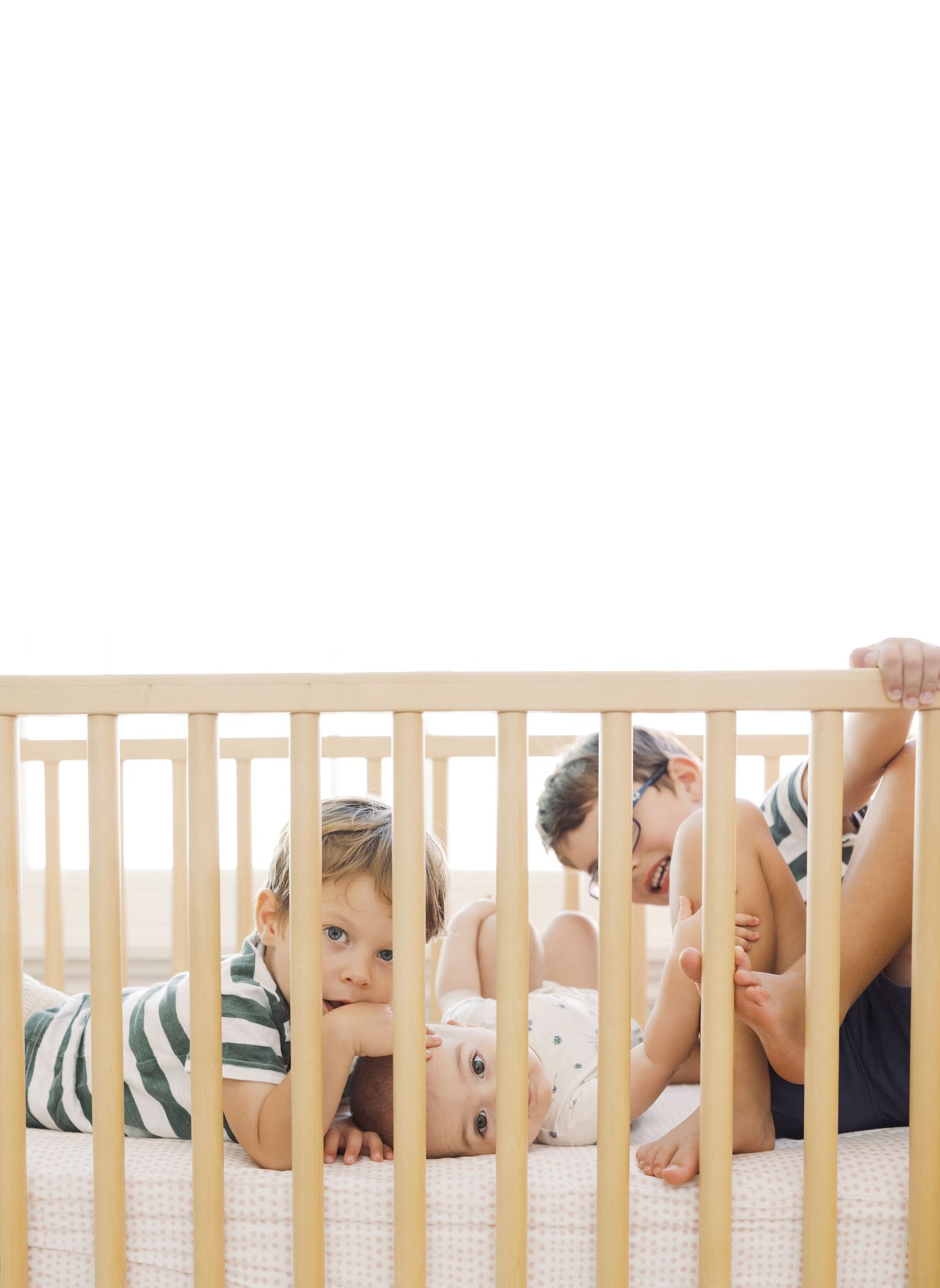 Two young children playing together inside a wooden baby crib with white bedding in a bright room.