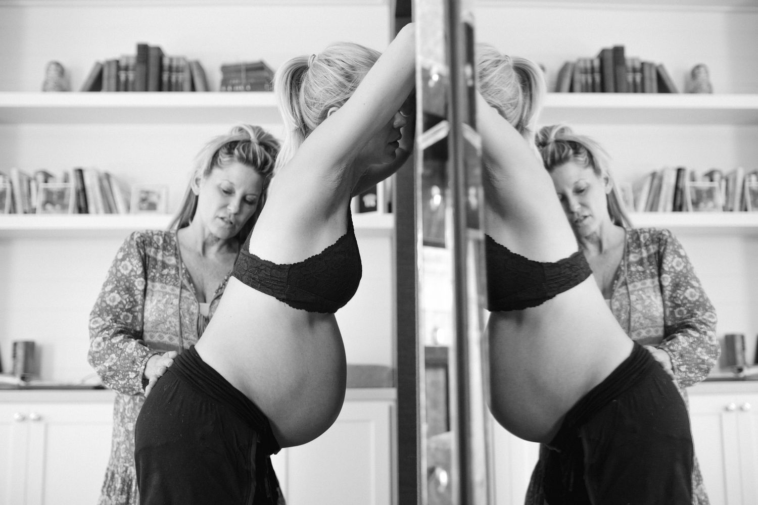 Black and white photo of a pregnant woman admiring her baby bump in a mirror at home.