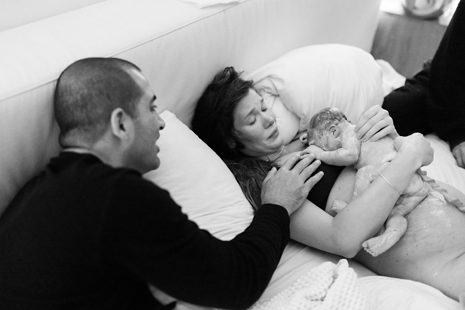 New mother lying in bed holds newborn baby while father looks on lovingly after home birth.