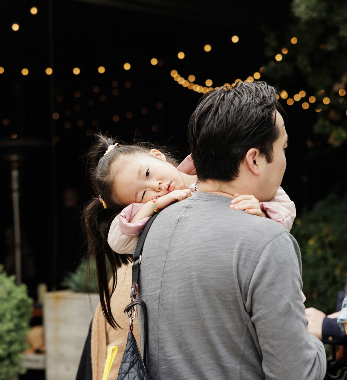 A young girl rests her head on her father's shoulder while being carried at an outdoor evening event.