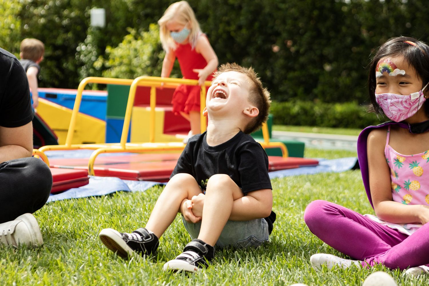 Joyful boy laughing while sitting on grass at an outdoor kids party with masked children nearby.