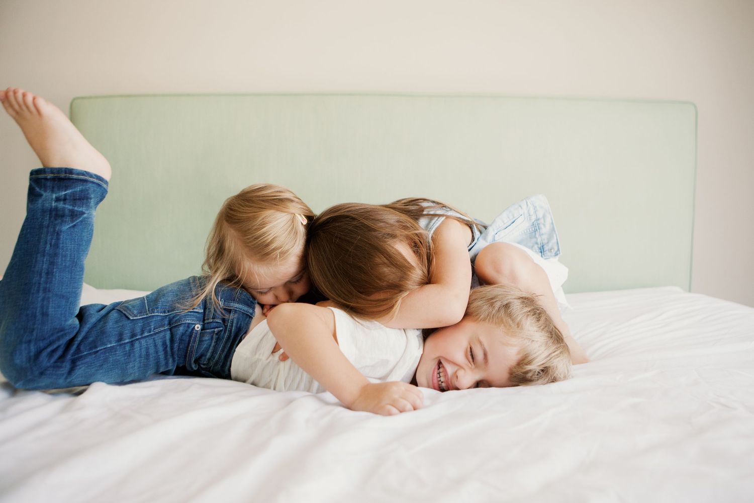 Three young children playfully pile on top of each other laughing on a white bed with a green headboard.