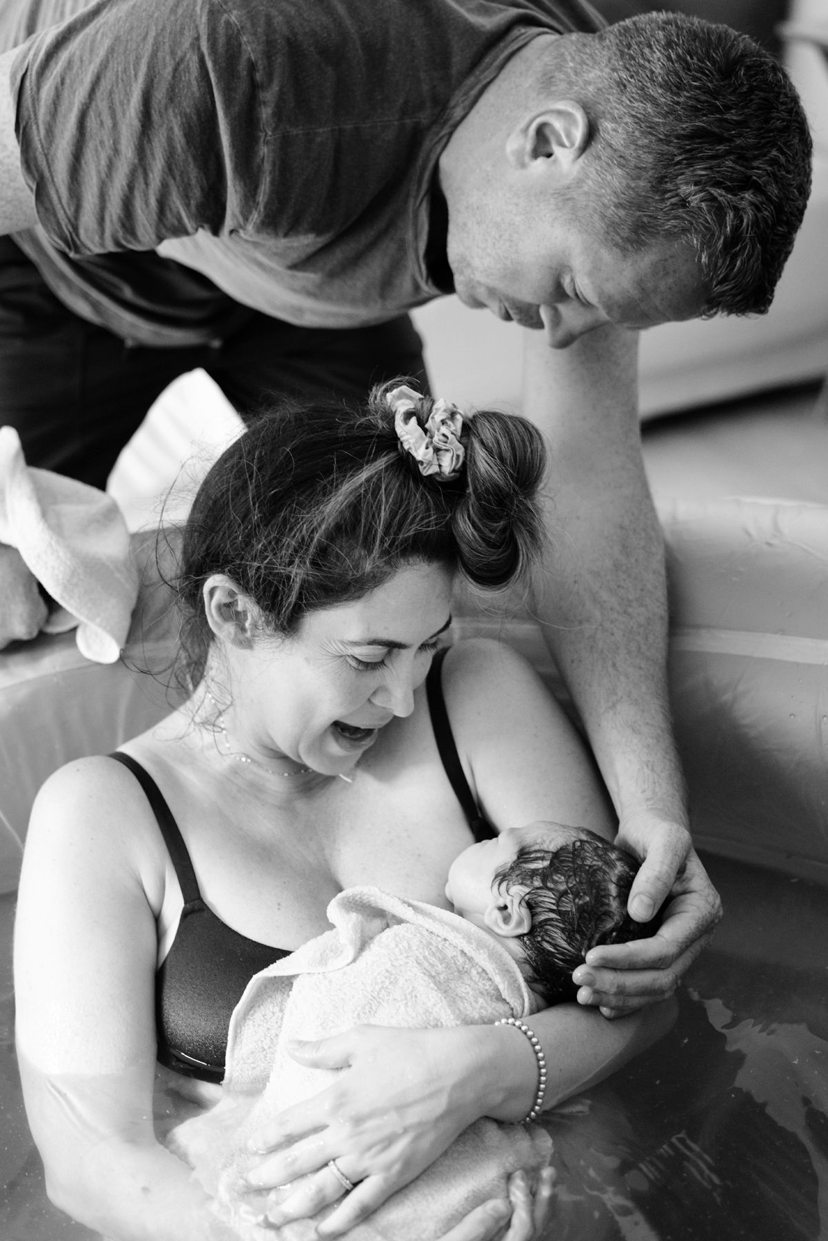 Emotional mother cradles newborn in water birth tub as father lovingly looks on after delivery.