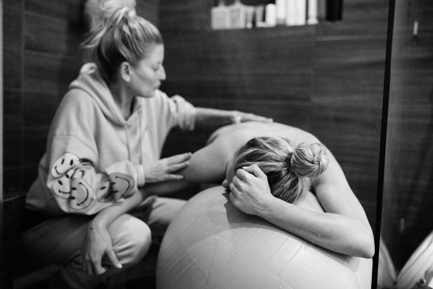 Doula providing back support to laboring woman leaning over birth ball.