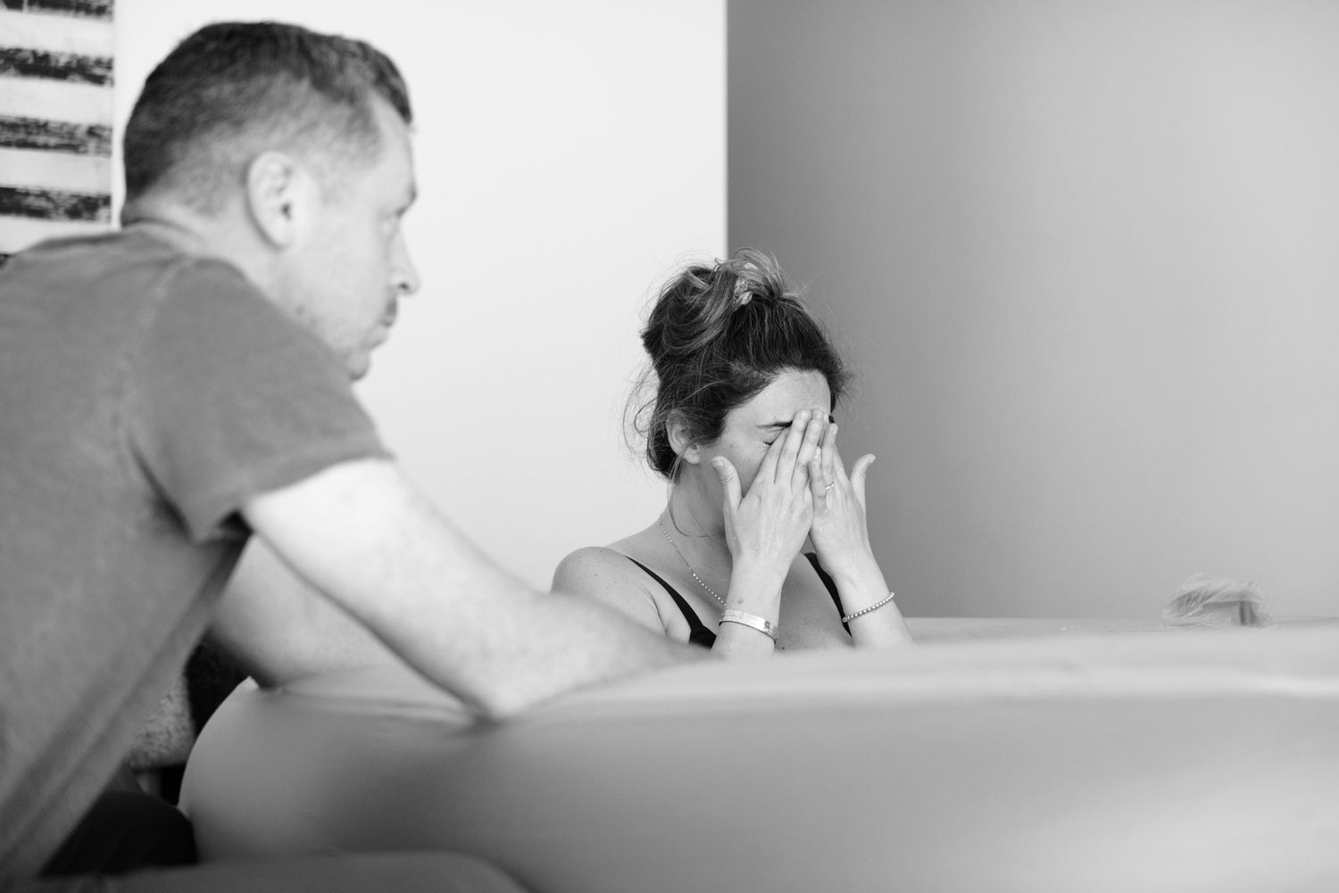 A woman covers her face during an intense labor contraction while her partner provides support beside a birthing tub.