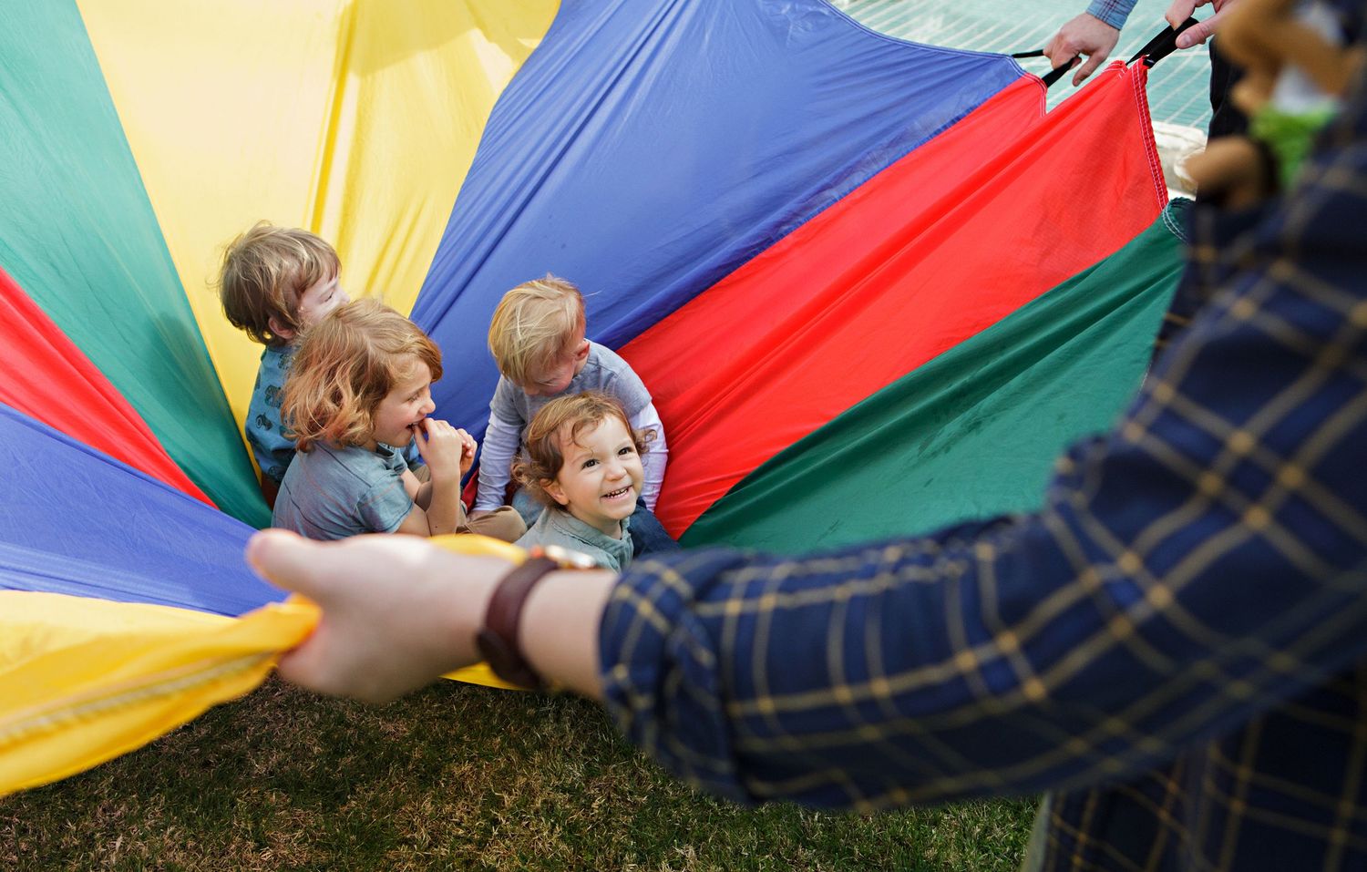Smiling toddlers peeking under a colorful parachute during an outdoor group play activity.