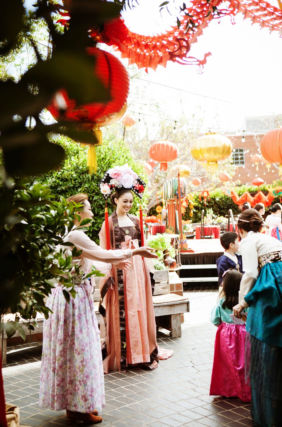 Women in elegant traditional Chinese hanfu dresses mingle outdoors at a Lunar New Year celebration with red lanterns.