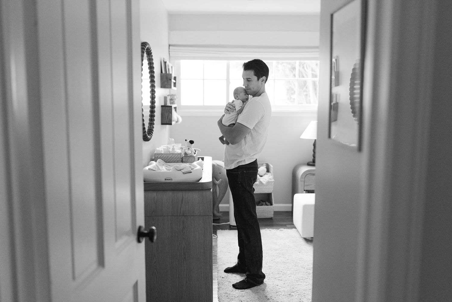 Black and white photo of a father cradling his newborn baby while standing in a nursery room.