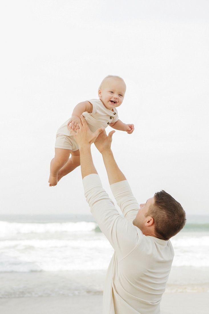 Father joyfully lifts smiling baby in the air on a sunny beach, both wearing white outfits with ocean waves behind them.