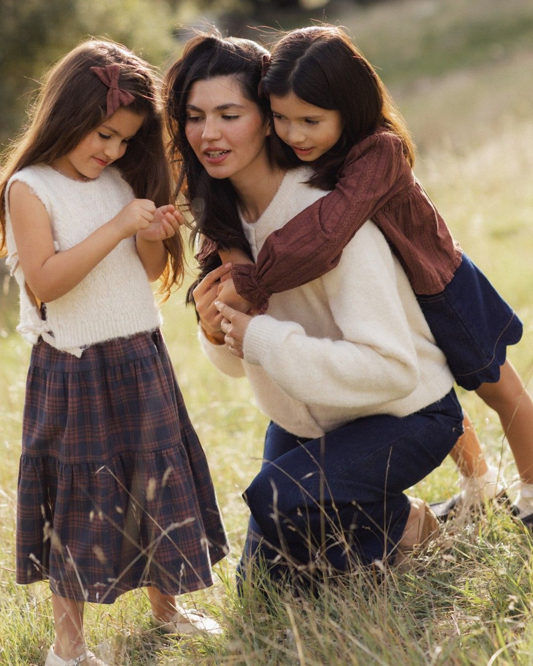 A mother kneels in a grassy field, hugging two young girls in plaid skirts and cozy sweaters.