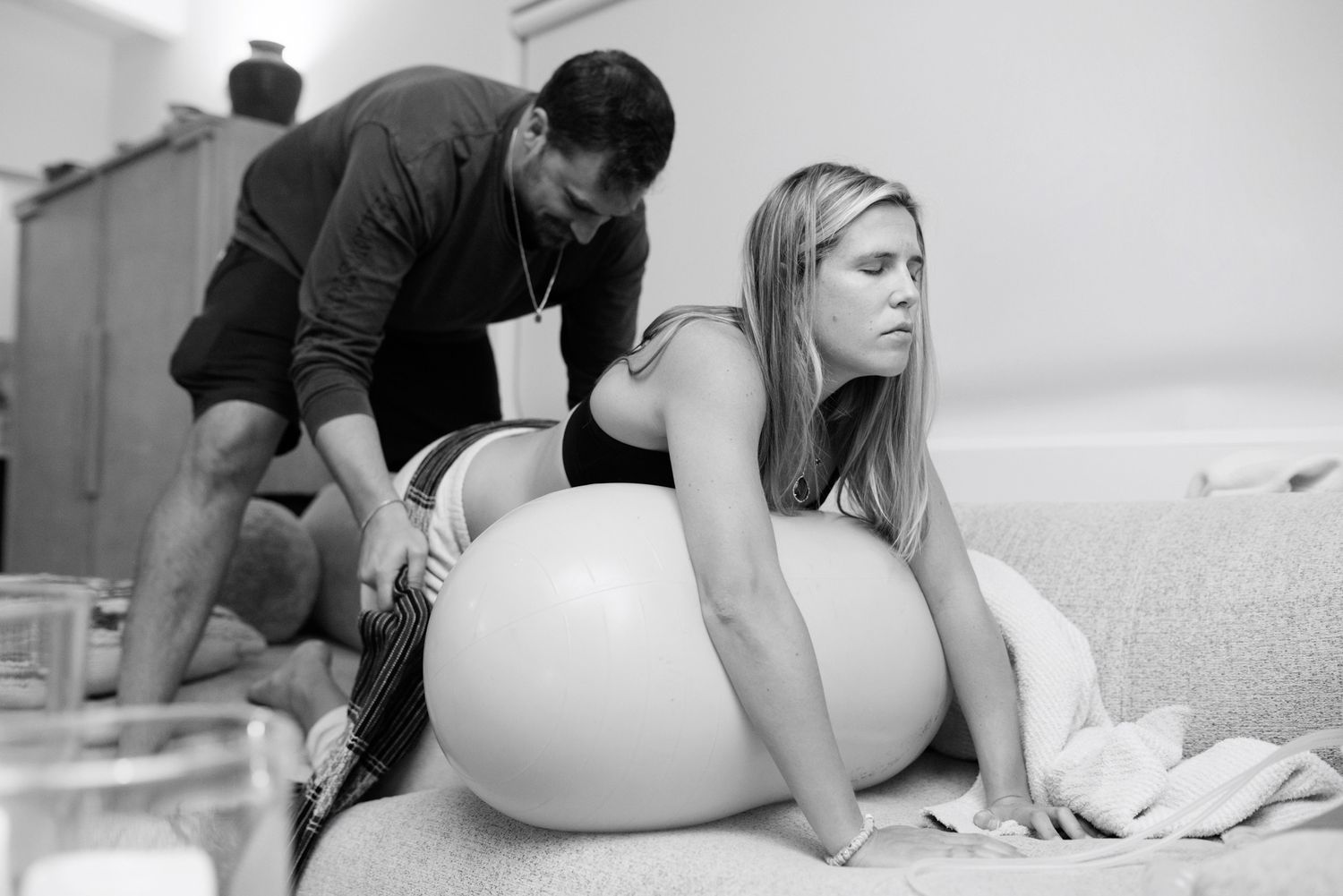 Partner supporting pregnant woman leaning on birth ball during labor at home.