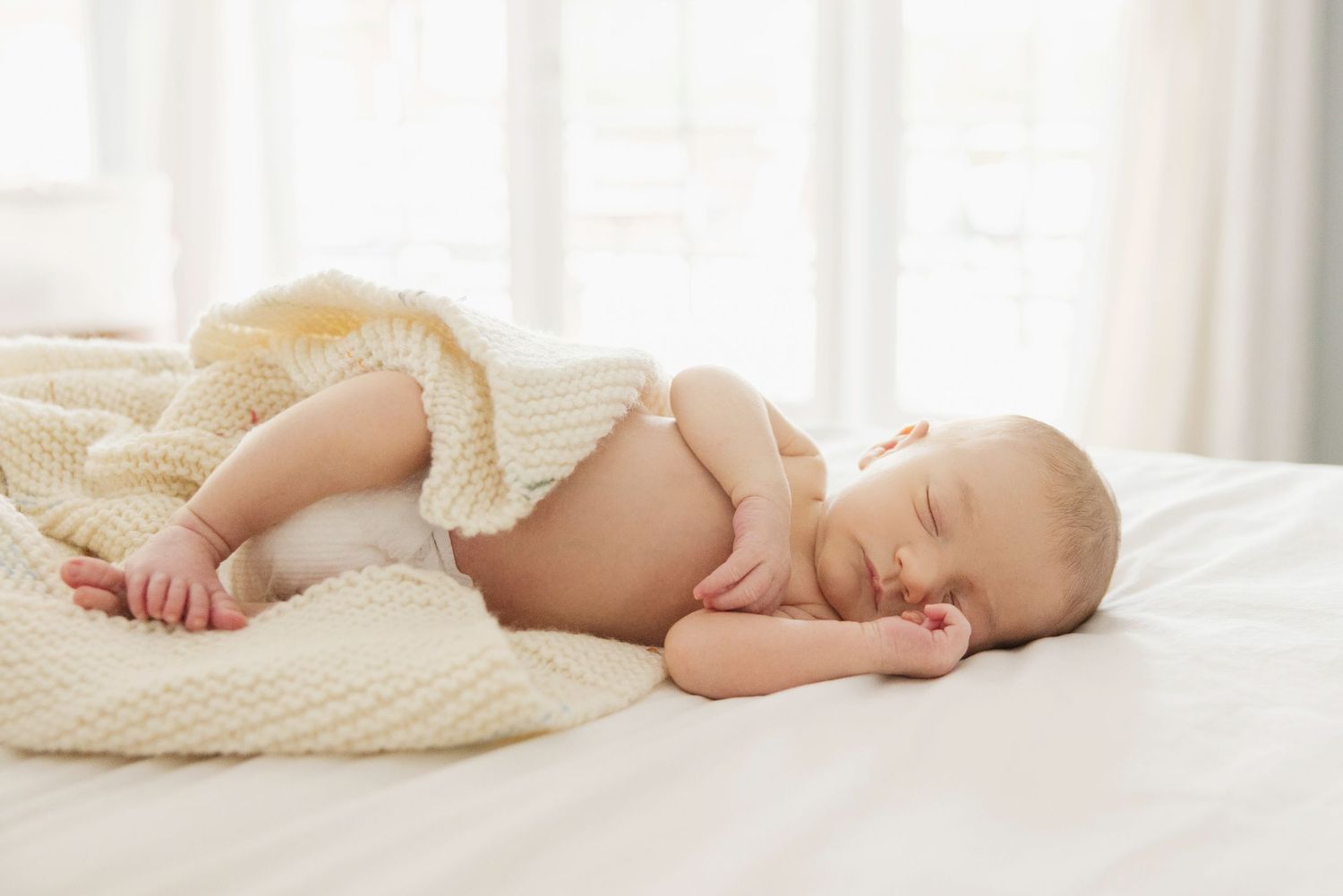 Sleeping newborn baby wrapped in cream knit blanket on white bed in bright room.