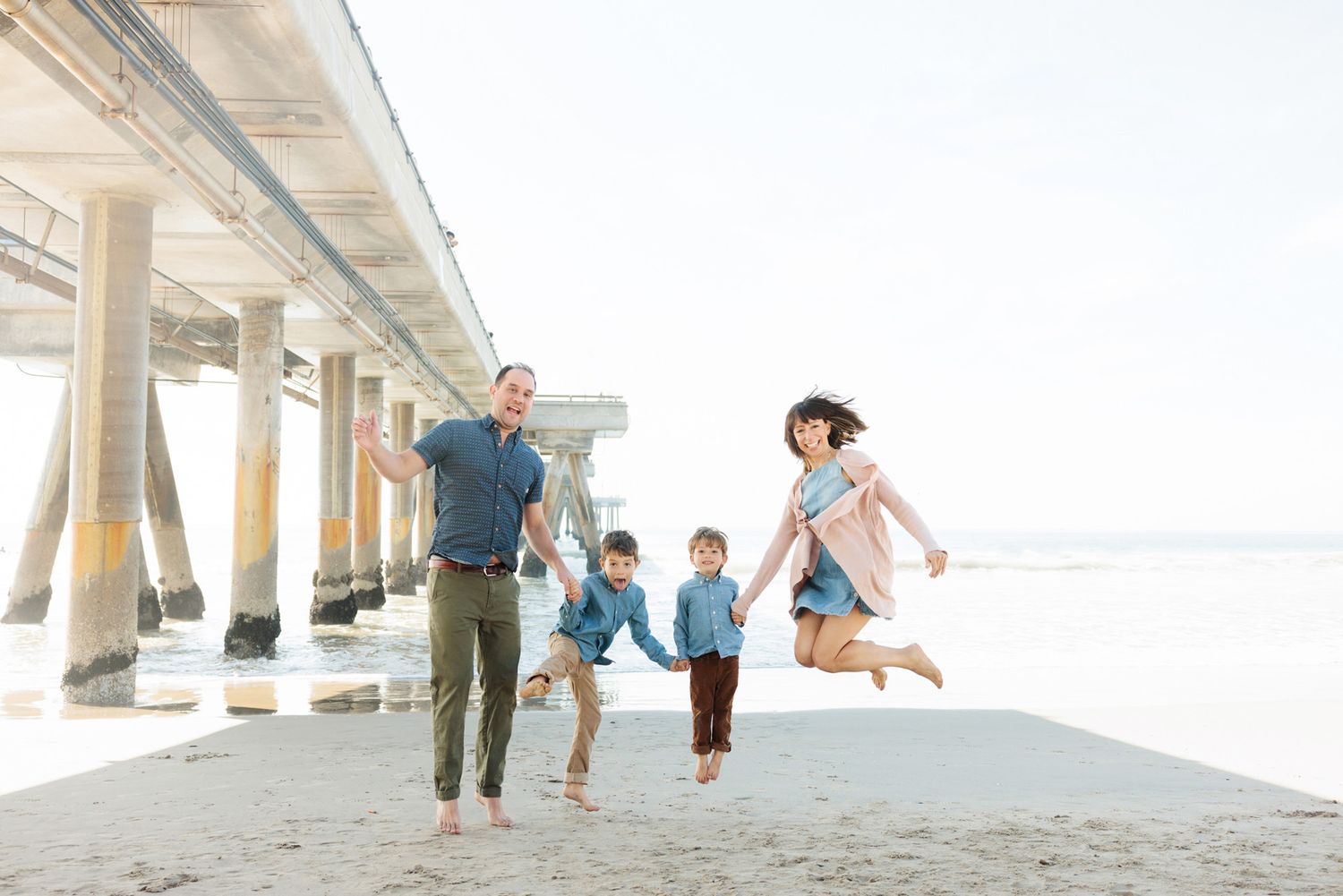 Family of four joyfully jumping and holding hands on a sandy beach near a pier on a bright day.