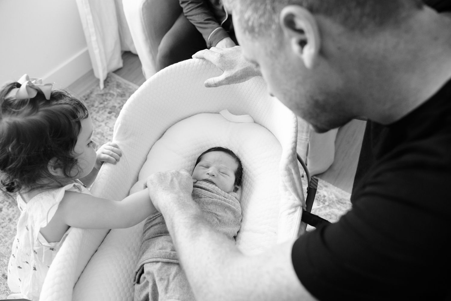 Father and young daughter lovingly admire newborn baby in white cushioned seat, black and white.
