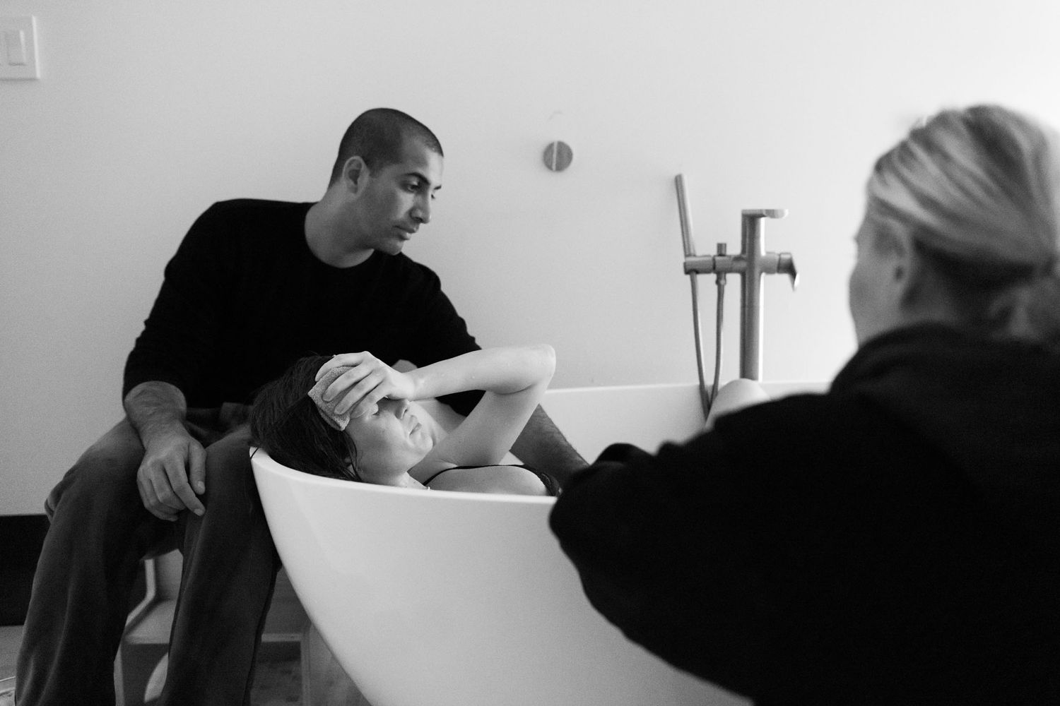 Man comforts laboring woman in a birthing tub as a midwife assists during a home water birth.