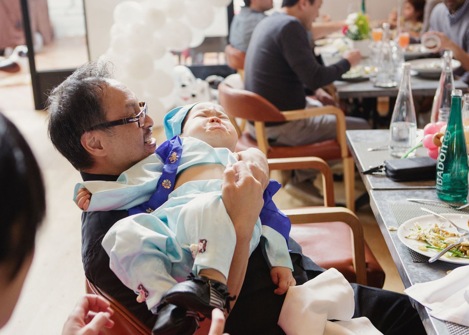 Father reclines with sleeping baby dressed in blue hanbok at restaurant birthday celebration table.
