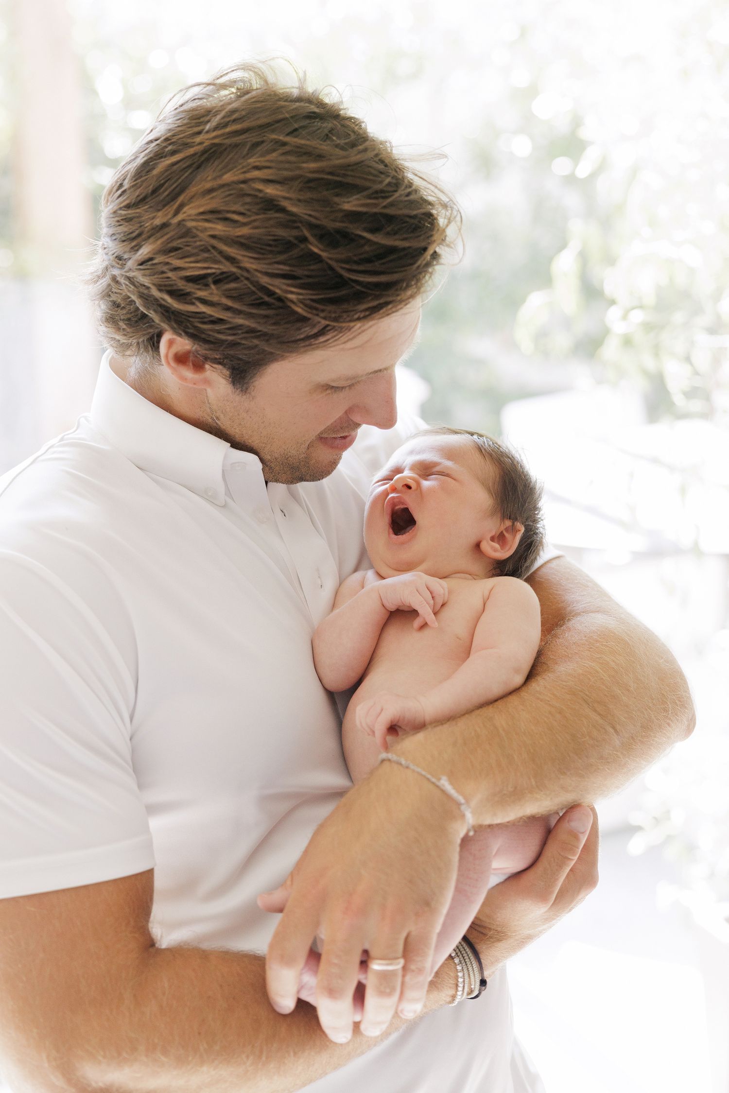 A father in a white polo shirt tenderly holds his yawning newborn baby close to his chest.