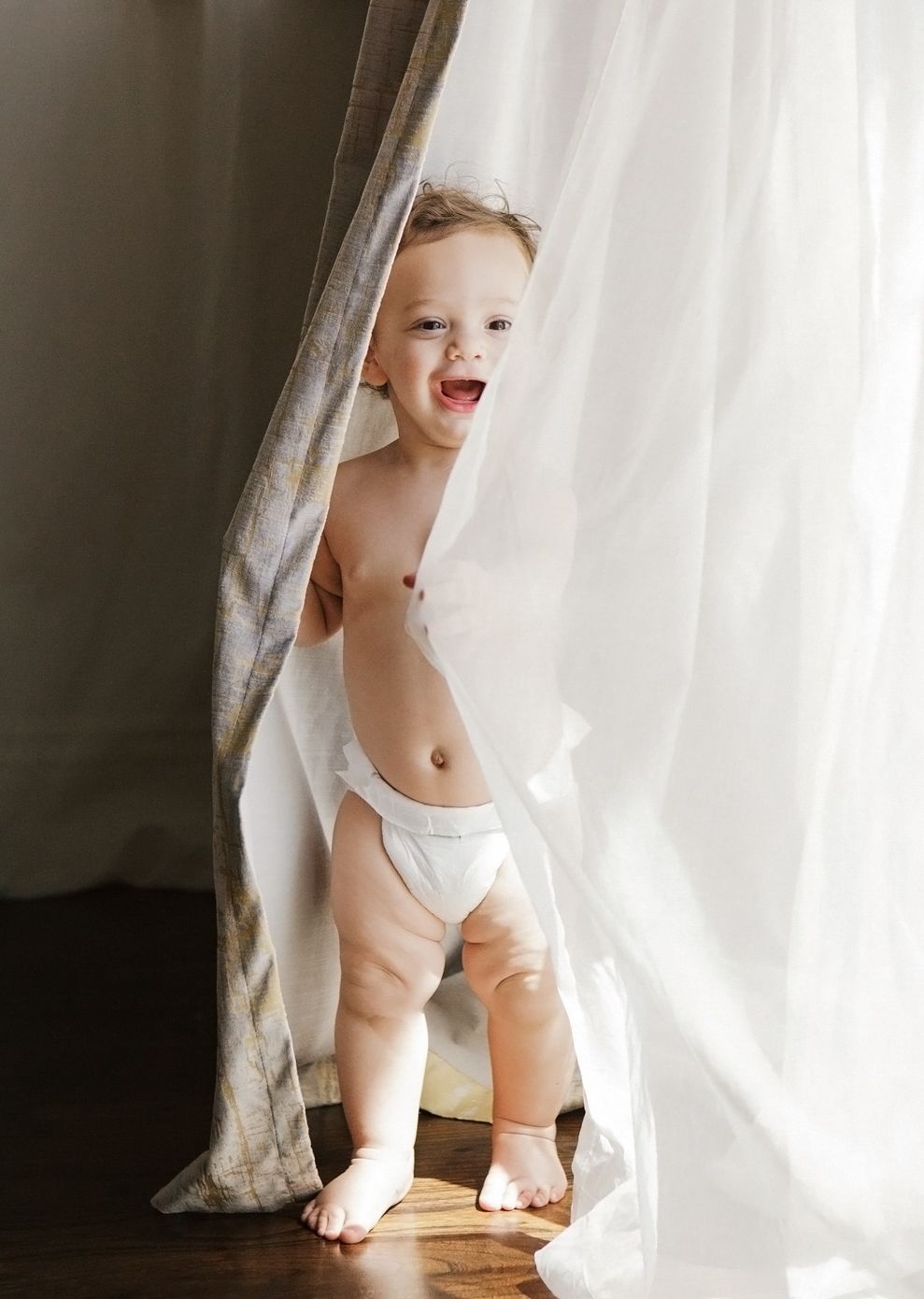 Happy toddler in diaper peeking through sheer white curtains, laughing and standing on hardwood floor.