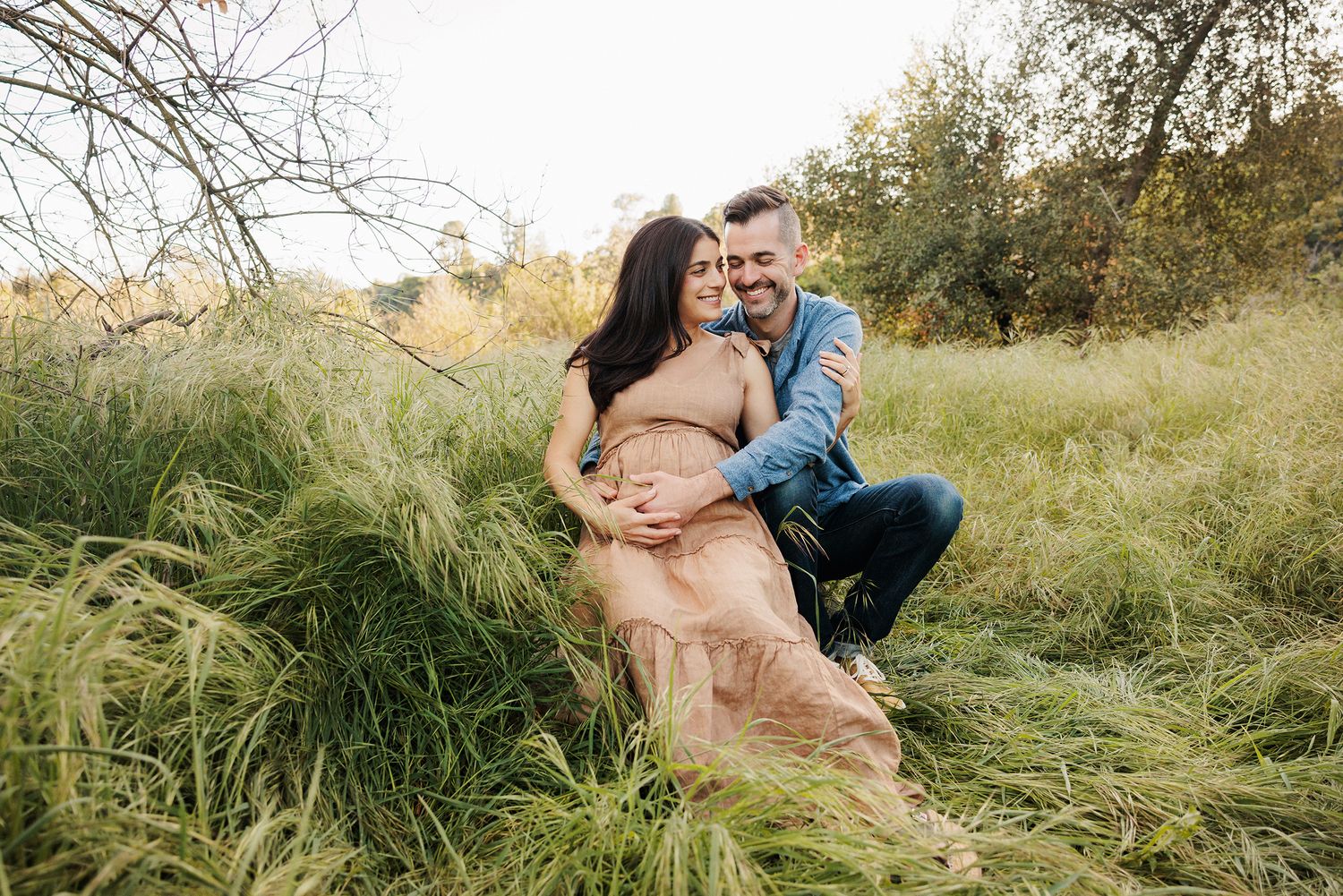 Couple embracing in lush green field during maternity photo session outdoors in autumn.