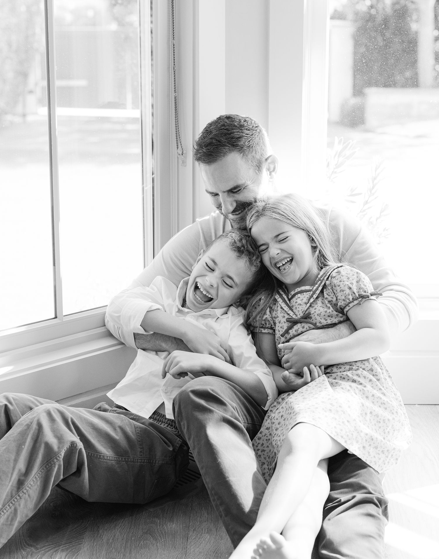 Black and white photo of a laughing father hugging two young children by a sunny window.