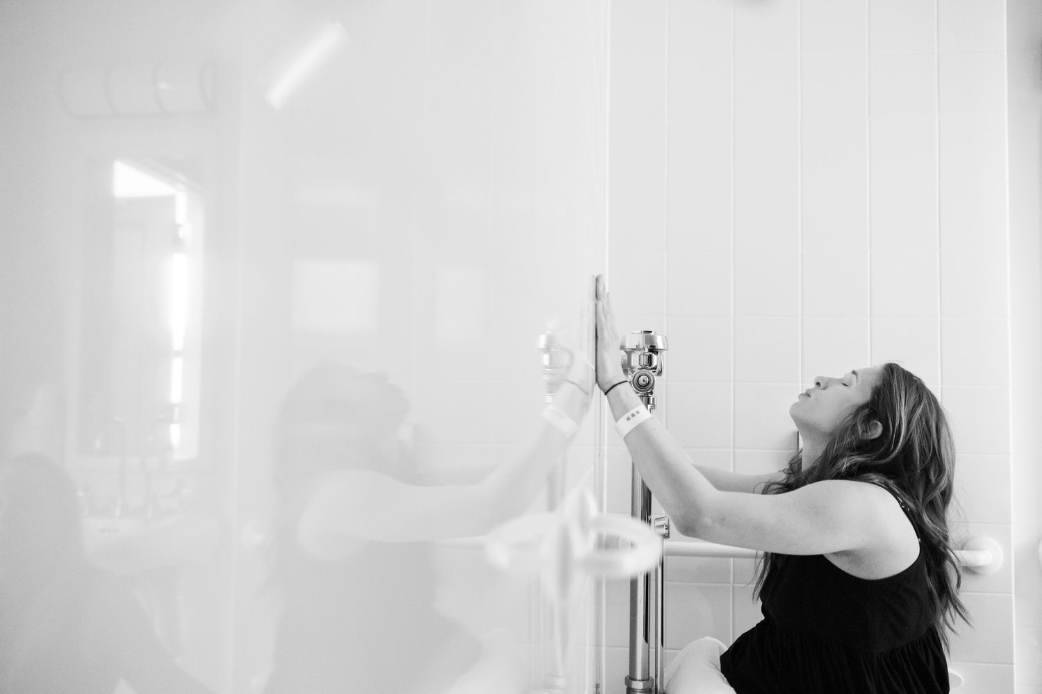 Black and white photo of a woman reaching up to adjust a faucet in a bright, minimalist bathroom.