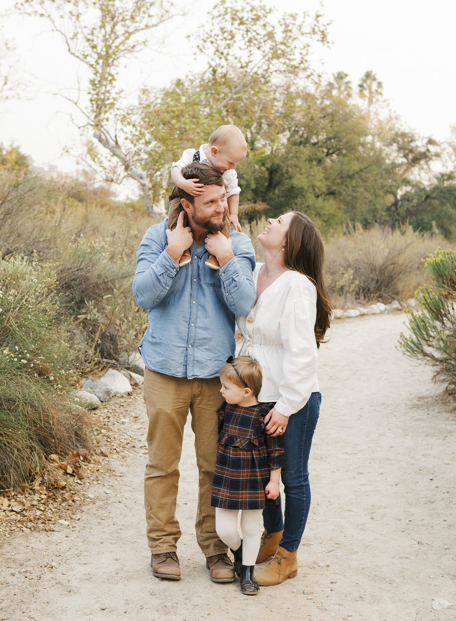 Family of four posing on a nature trail, parents smiling at baby held overhead, young girl standing beside them.