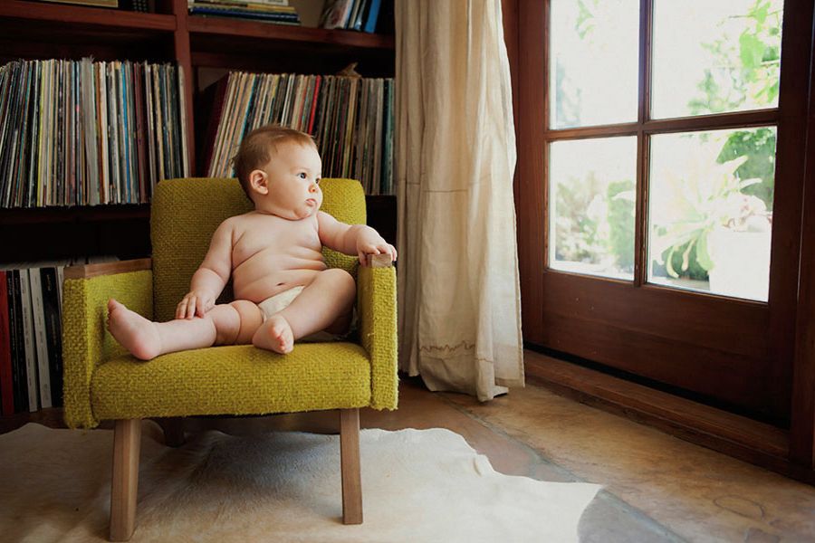A curious baby sits in a small green armchair next to a bookshelf of vinyl records and a sunlit window.