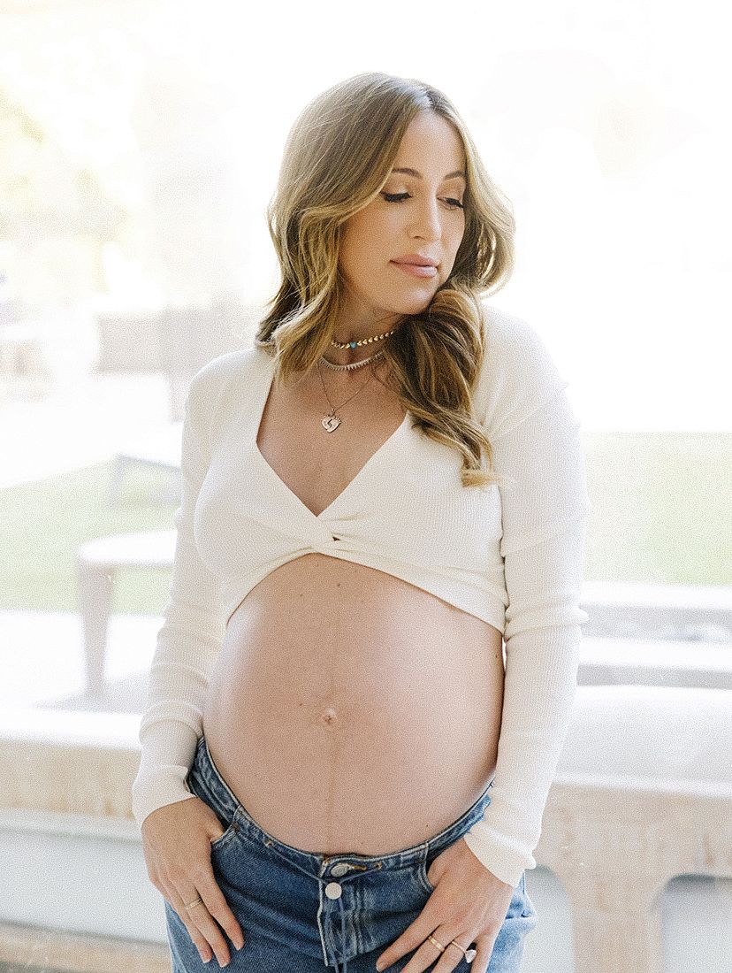 Pregnant woman in white crop top and jeans showing baby bump, wearing layered necklaces, looking down softly outdoors.