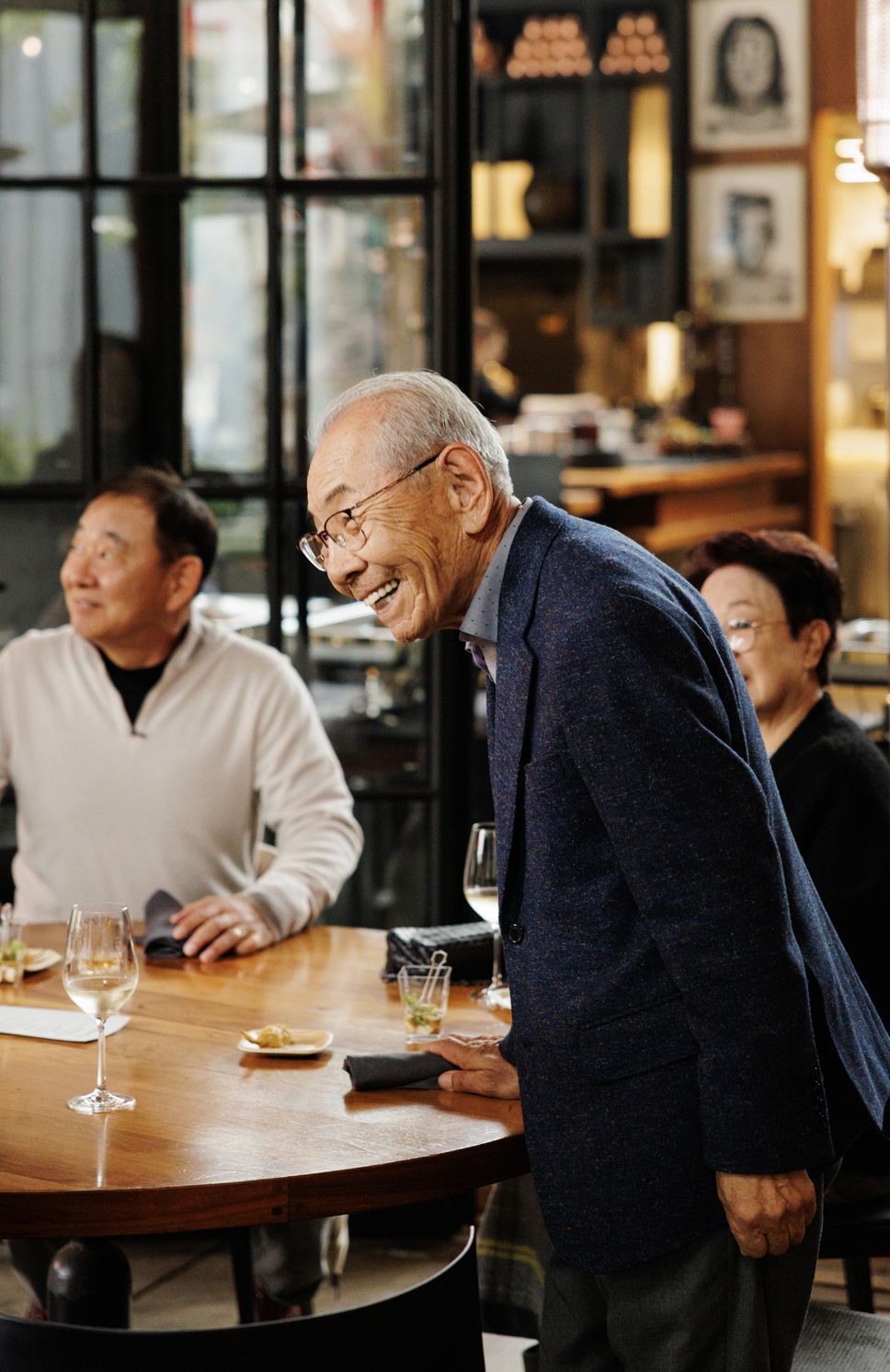 A smiling elderly Asian man in a navy blazer laughs while leaning on a wooden table at a restaurant gathering.