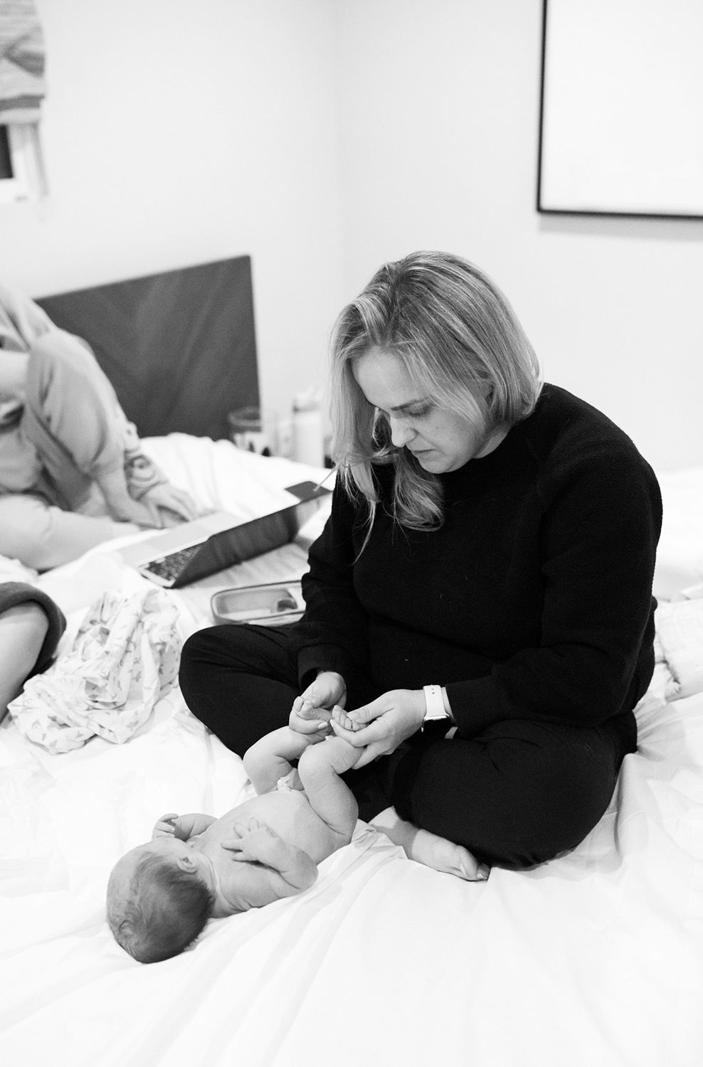 Woman in black outfit tends to newborn baby lying on white bed, intimate black and white photography.