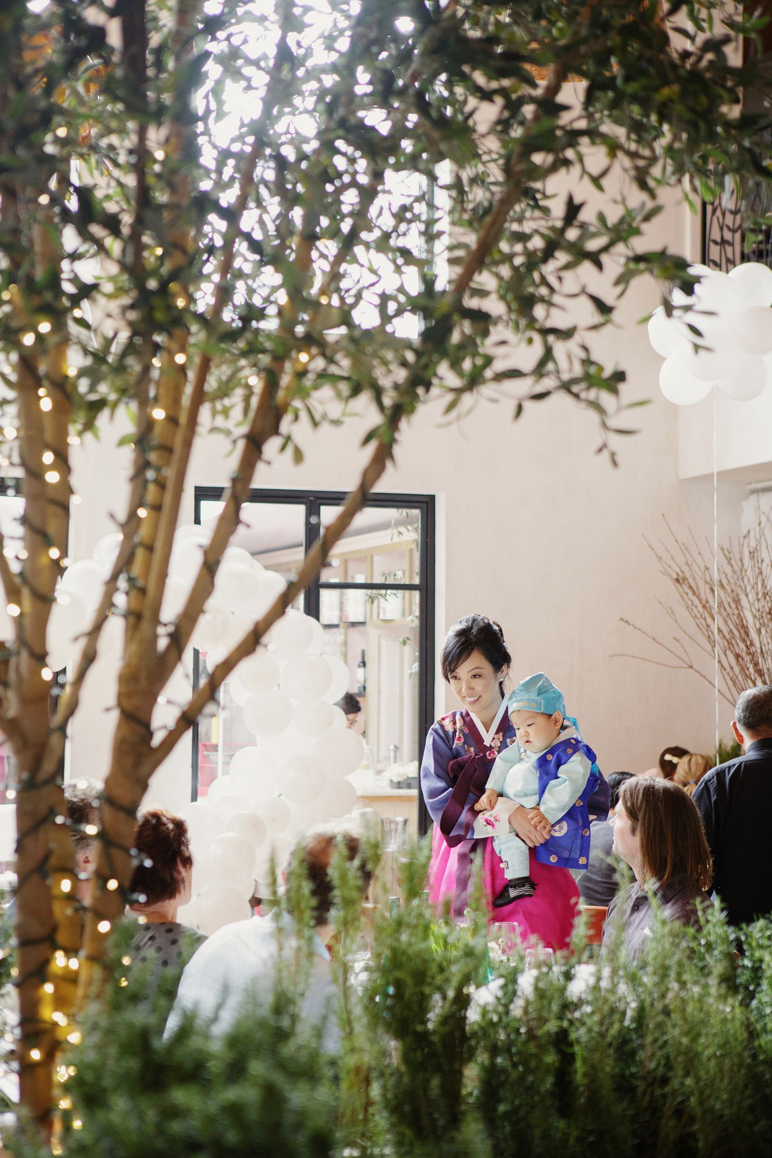 Mother in pink Korean hanbok holds baby in blue hanbok at decorated indoor garden party venue.