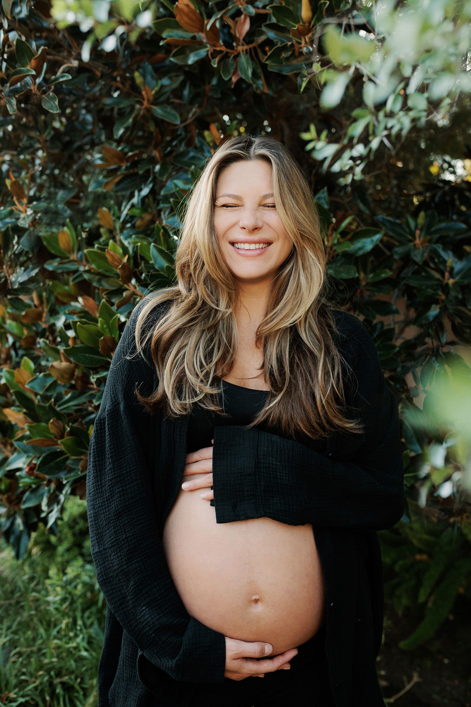 Smiling pregnant woman in black outfit holding baby bump outdoors near lush green magnolia leaves.