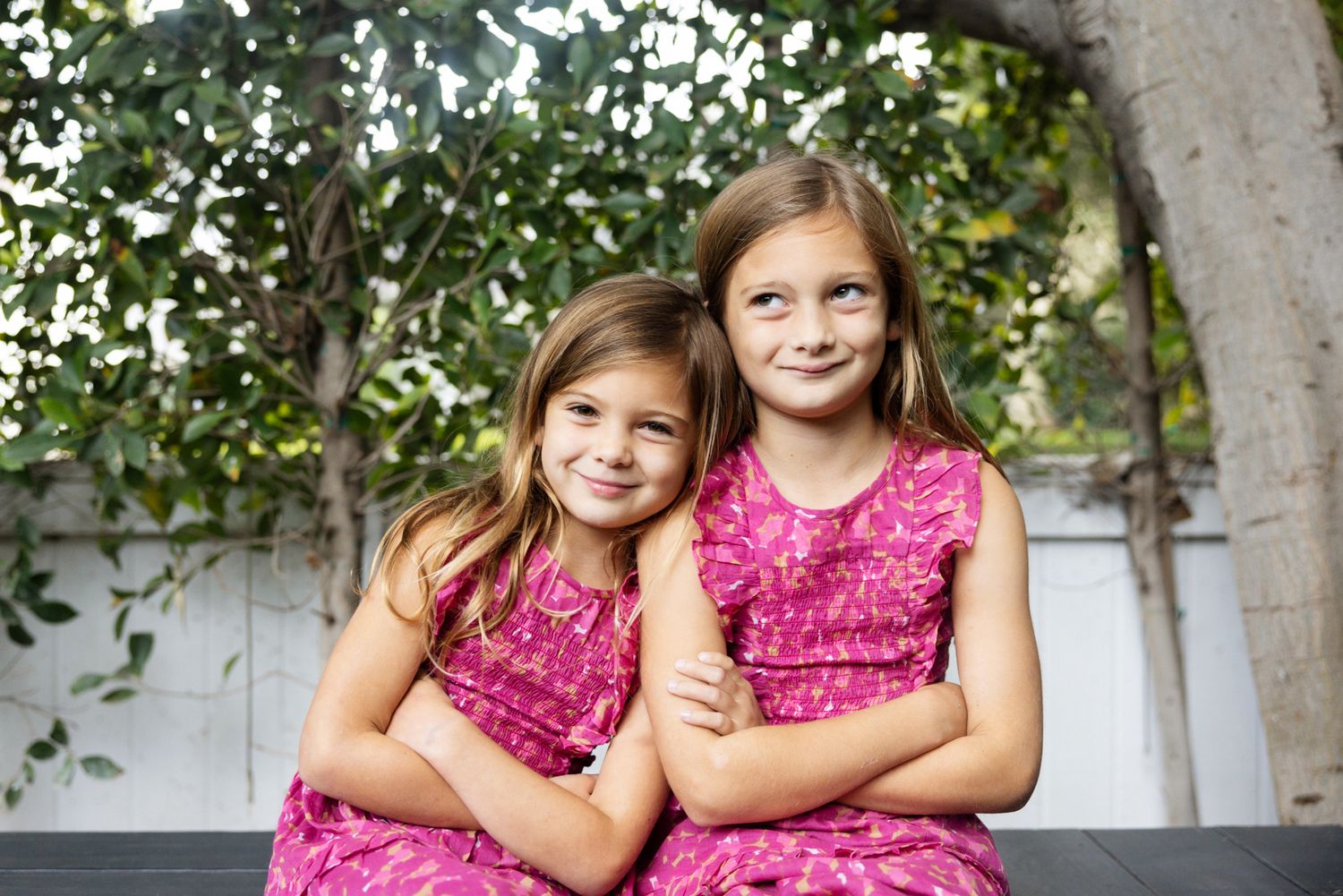 Two young sisters in matching pink floral dresses pose together outdoors under a green tree.