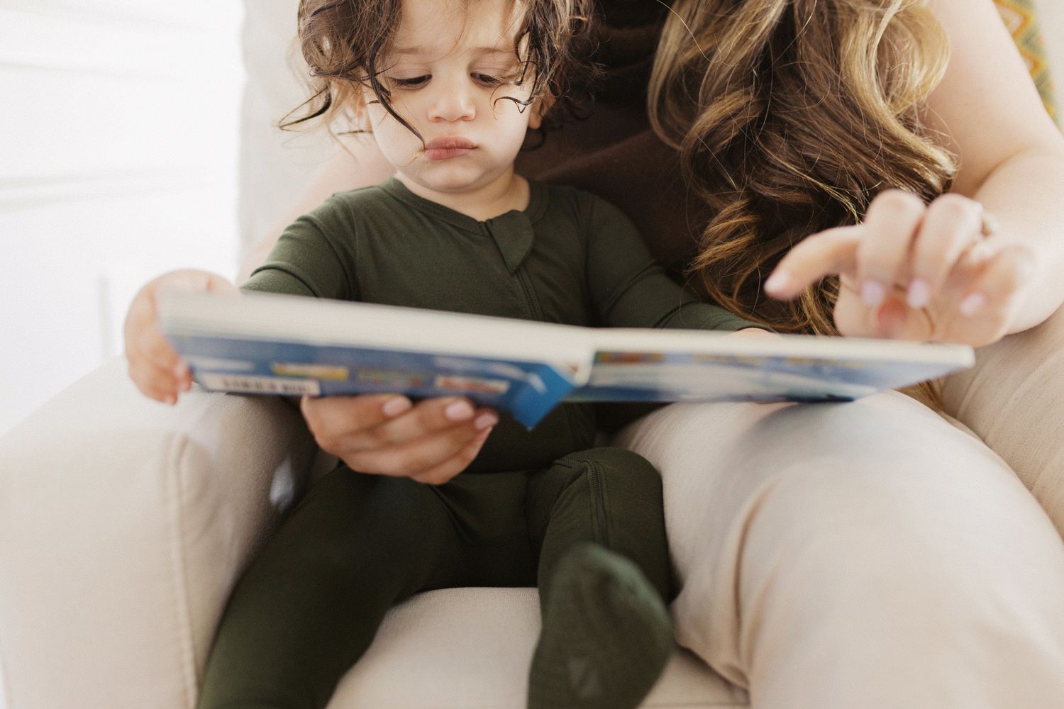 A young toddler in green pajamas sits on a lap reading a colorful picture book with an adult.