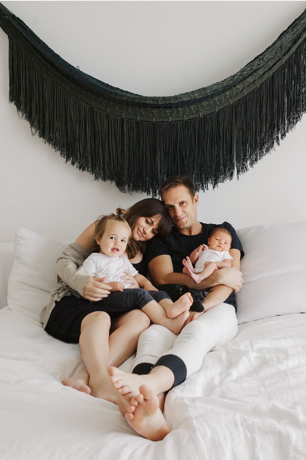 A young family of four sits on a white bed, parents holding a toddler and newborn beneath a black fringe wall hanging.