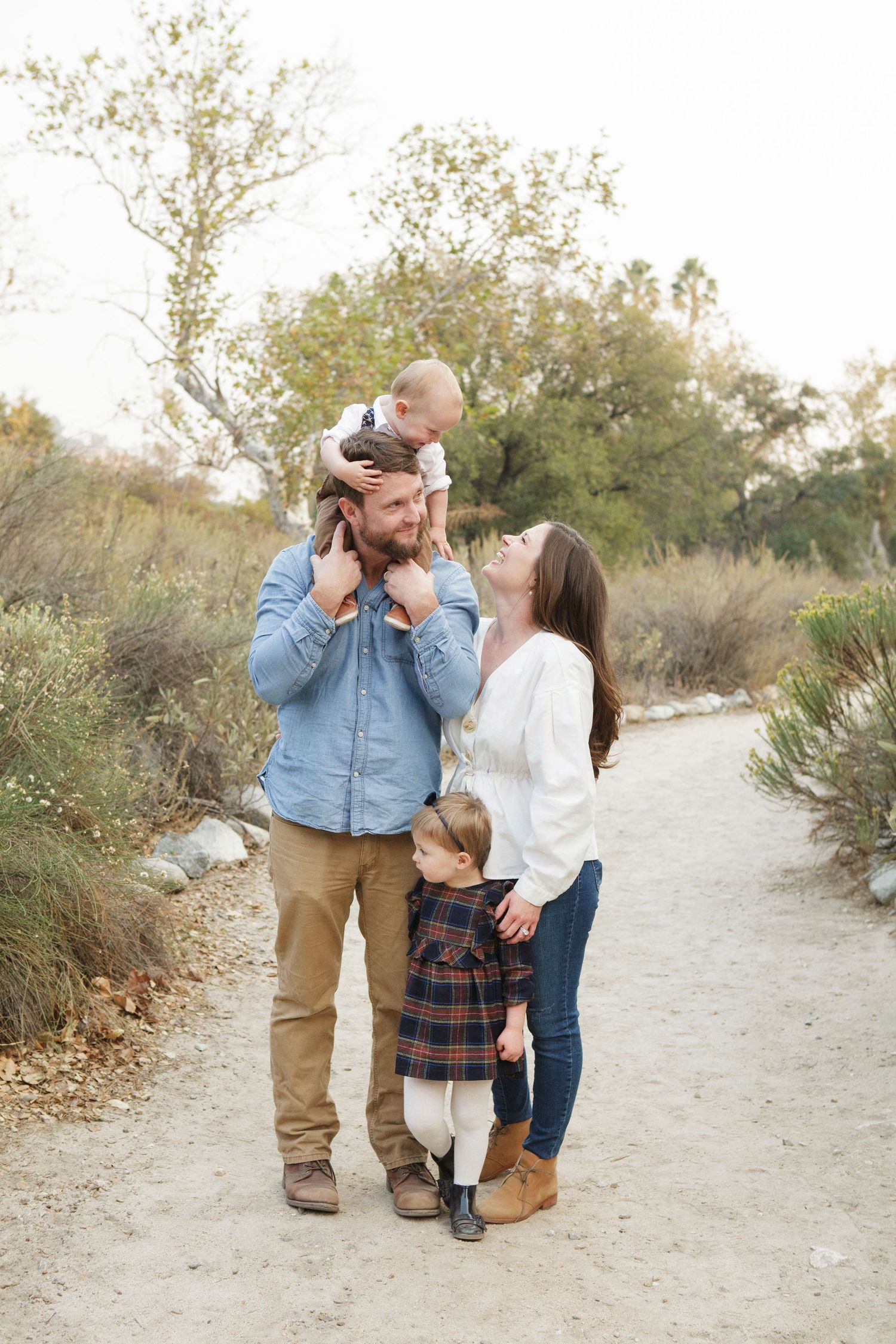 Family of four posing on a nature trail, father lifting baby overhead while mother and toddler stand beside him.