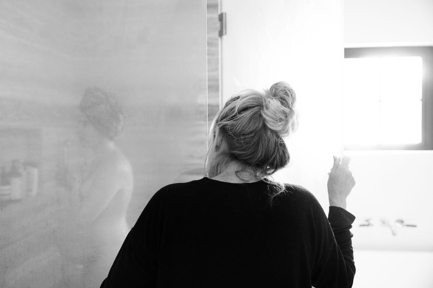 Blonde woman with hair in bun standing at bathroom mirror during home birth.