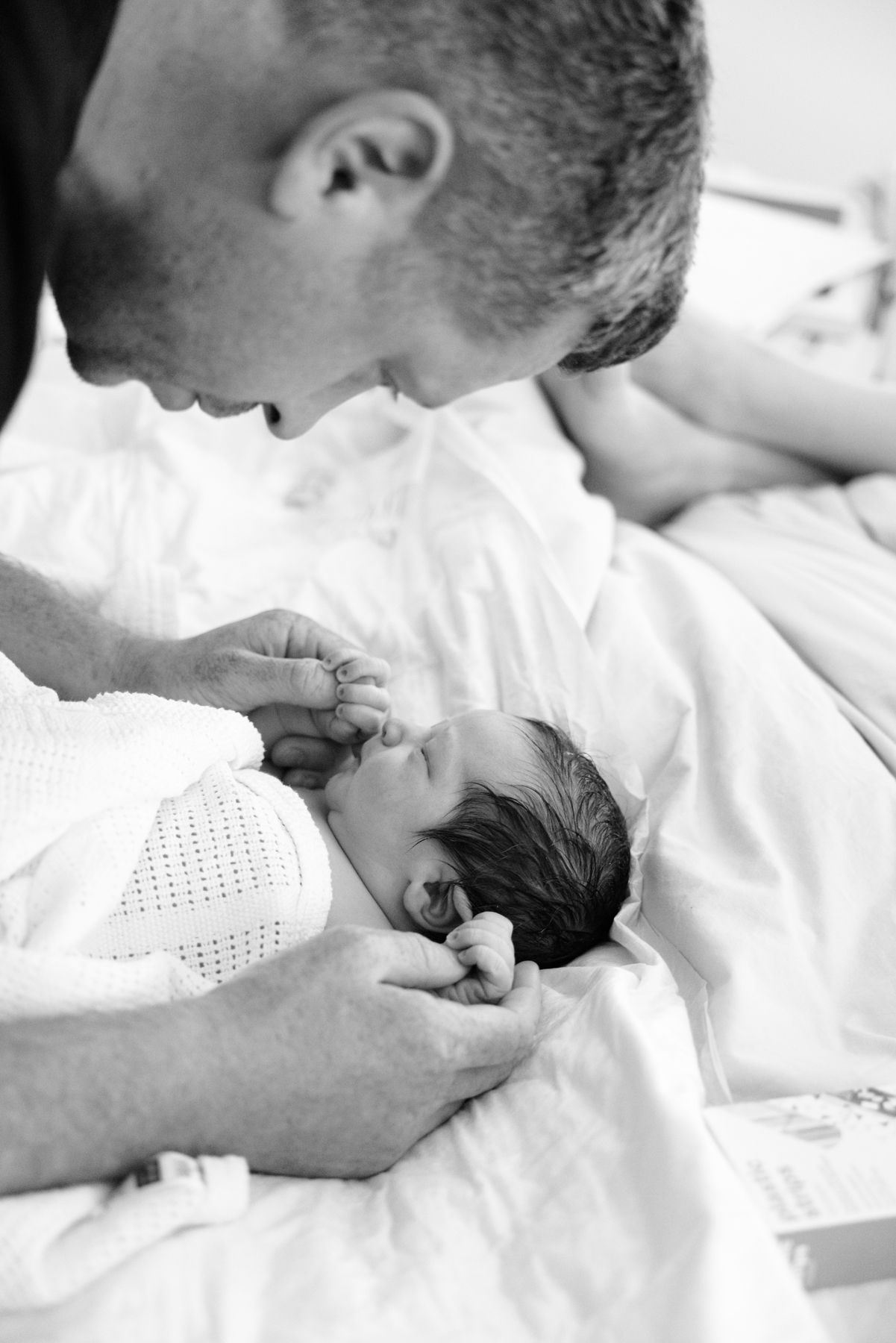 Father tenderly touches newborn baby's face while cradling infant in hospital bed, black and white photography.