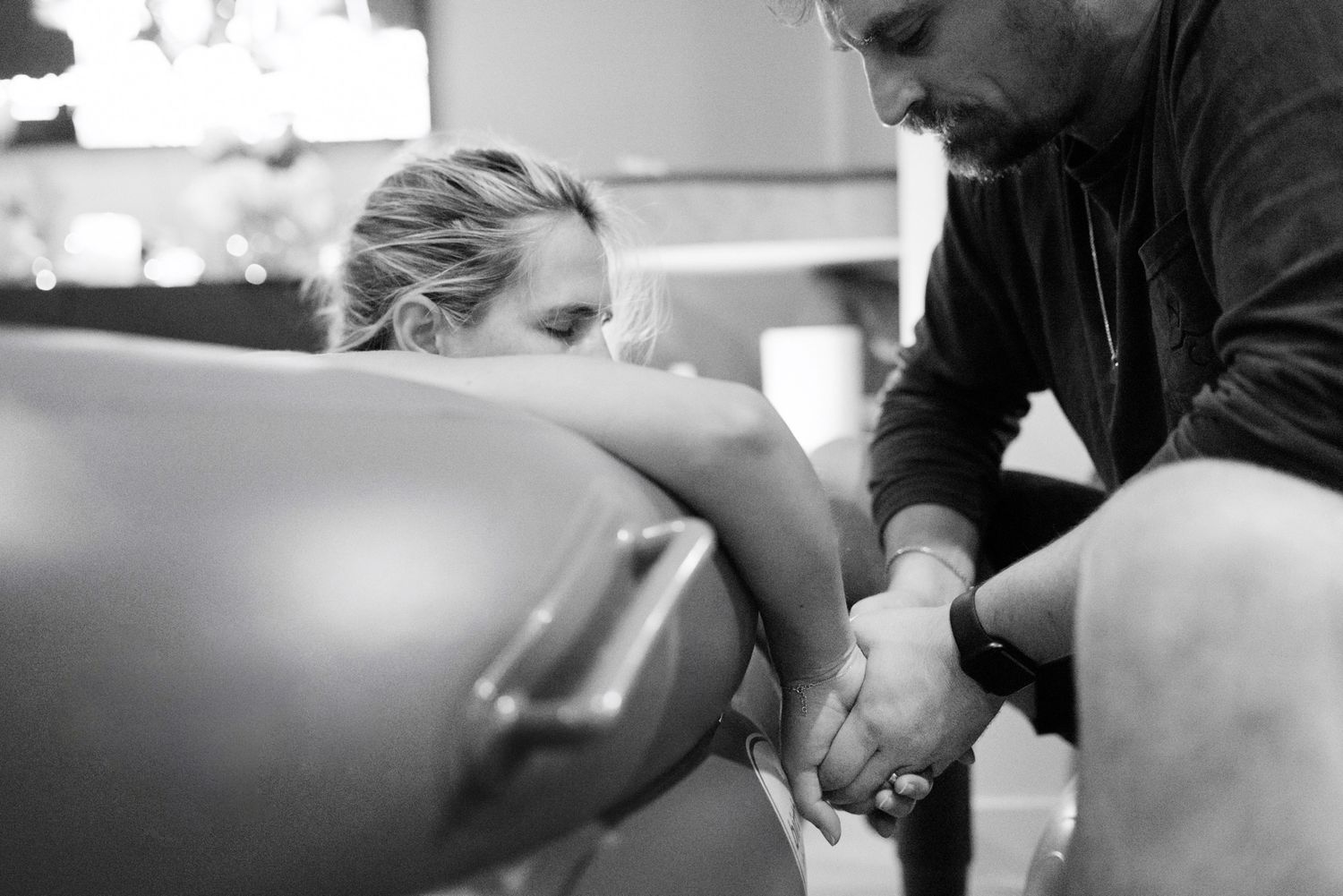 Black and white image of a bearded man supporting a woman during water birth labor, holding hands beside birth pool.