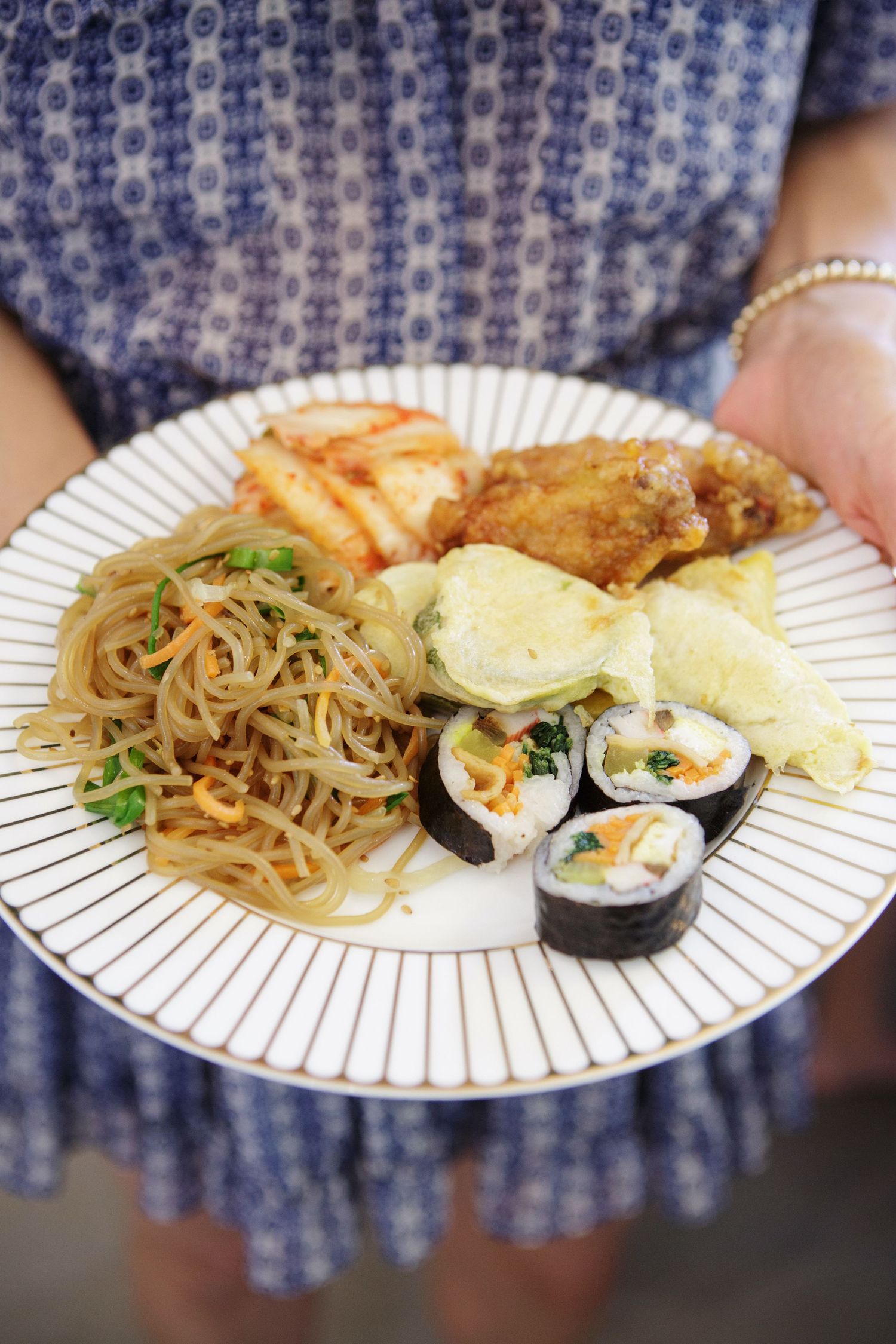 Person holding plate of Korean food including japchae noodles, kimbap rolls and fried items.