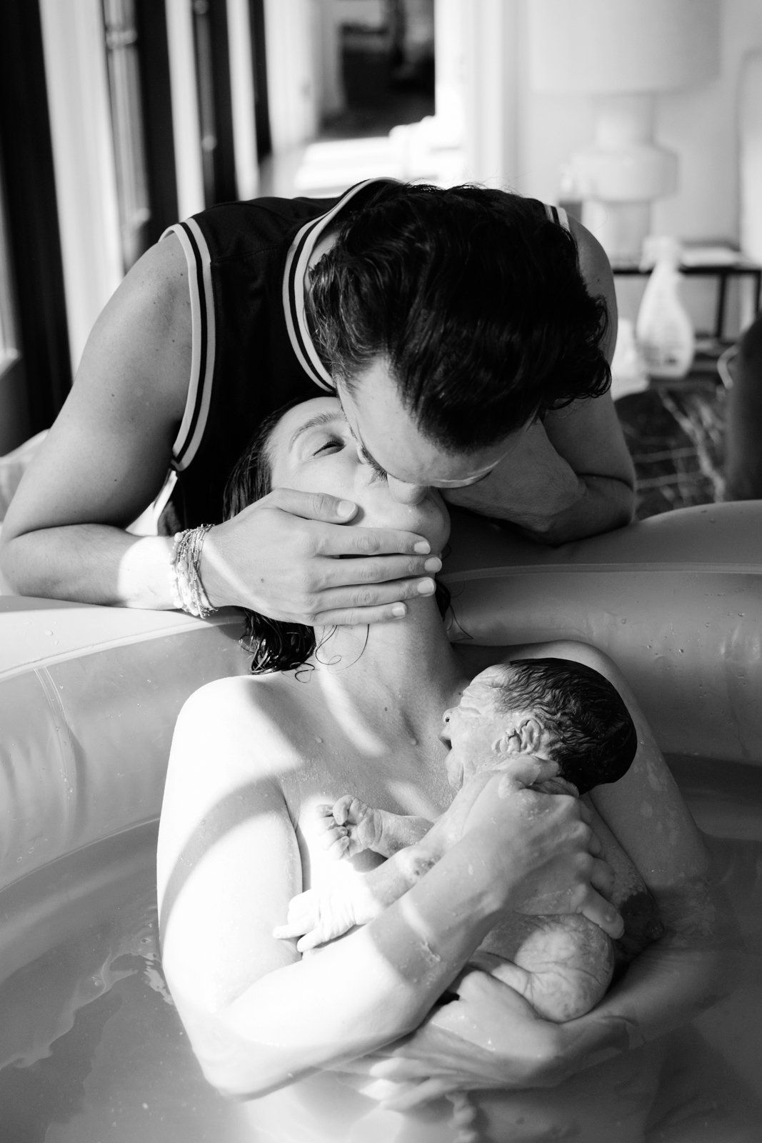 Black and white photo of a partner kissing a mother holding her newborn baby in a water birth tub.