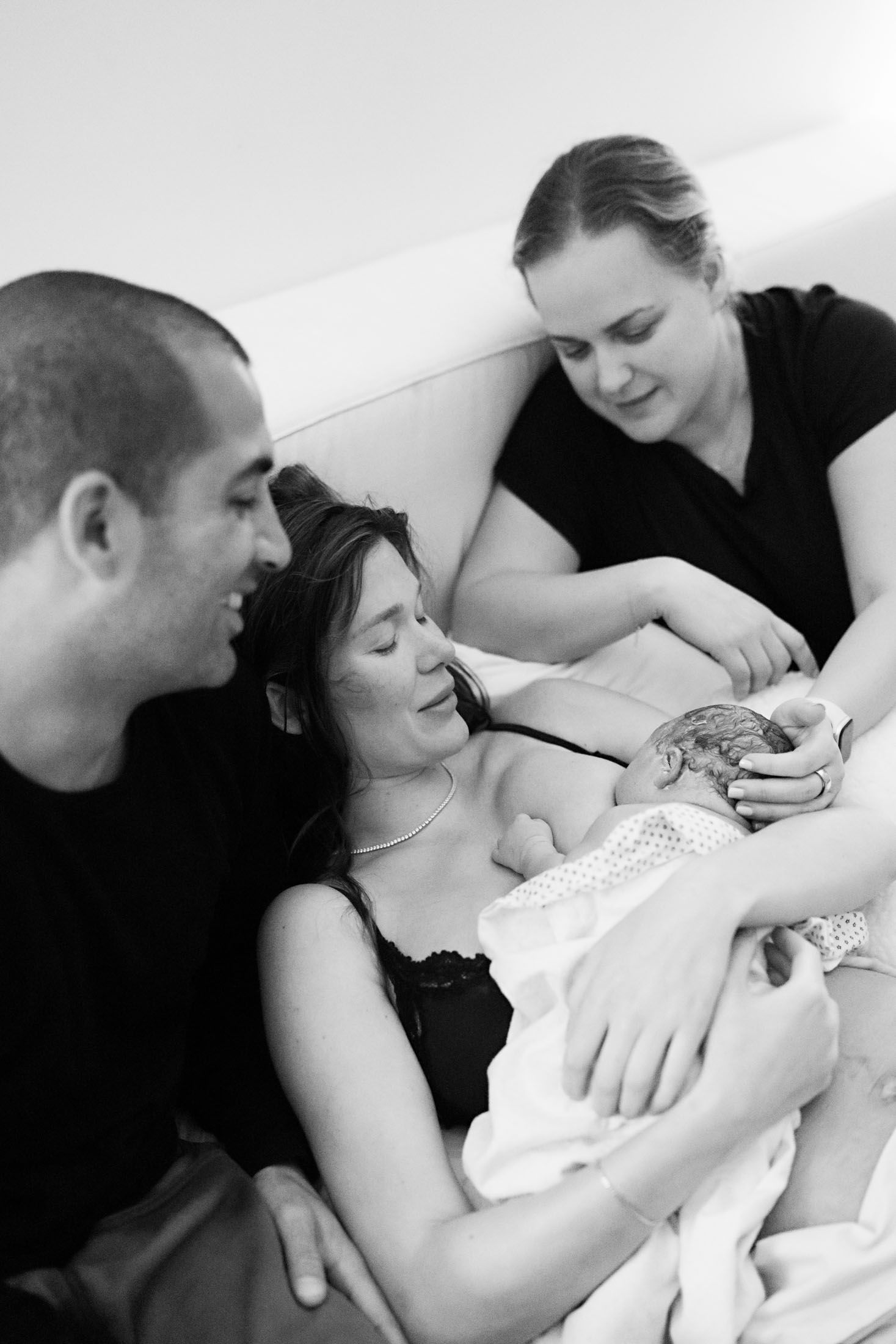 Father and midwife support smiling mother holding newborn baby after home birth delivery.