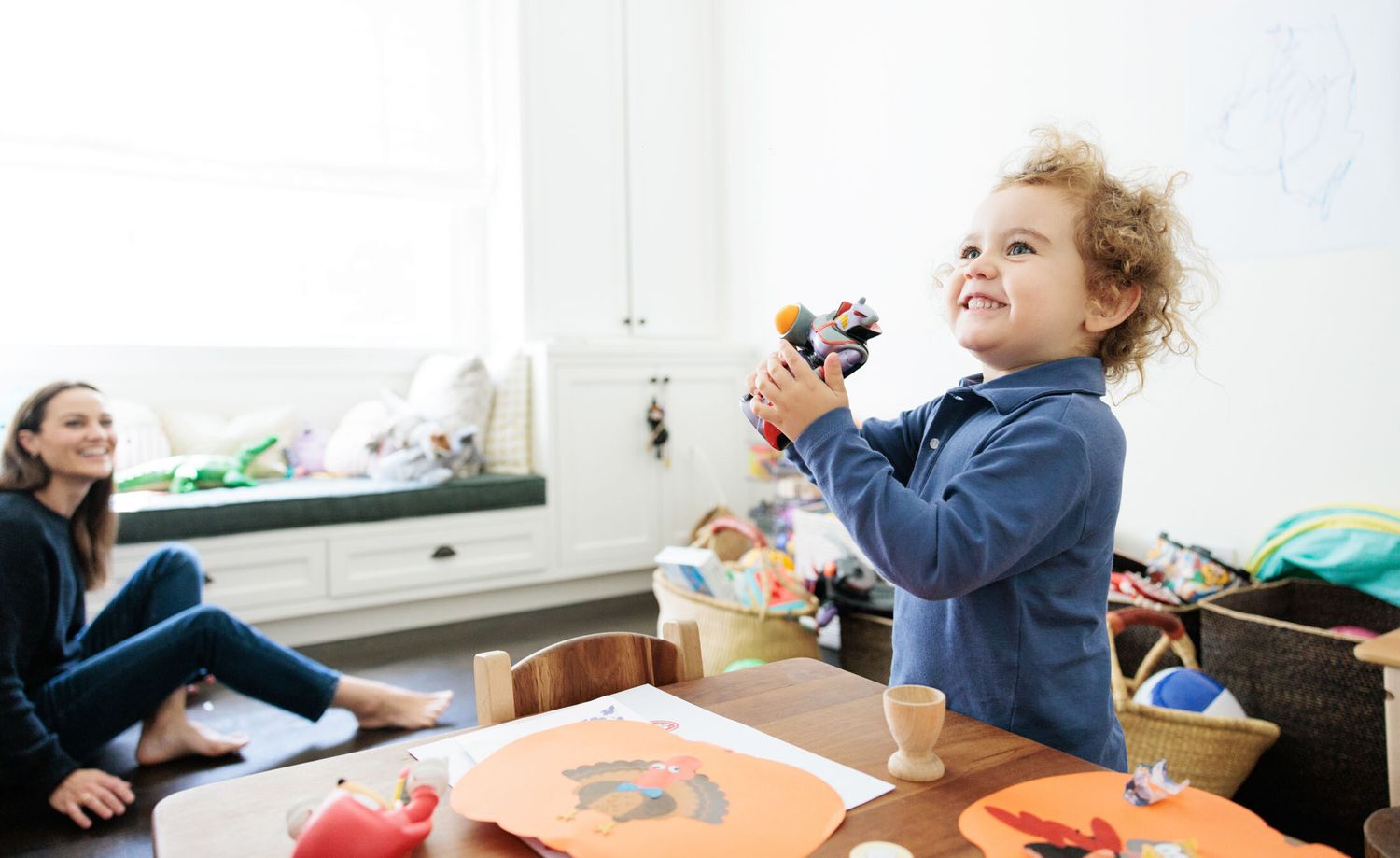 Happy toddler playing with toy while mom watches in bright playroom.