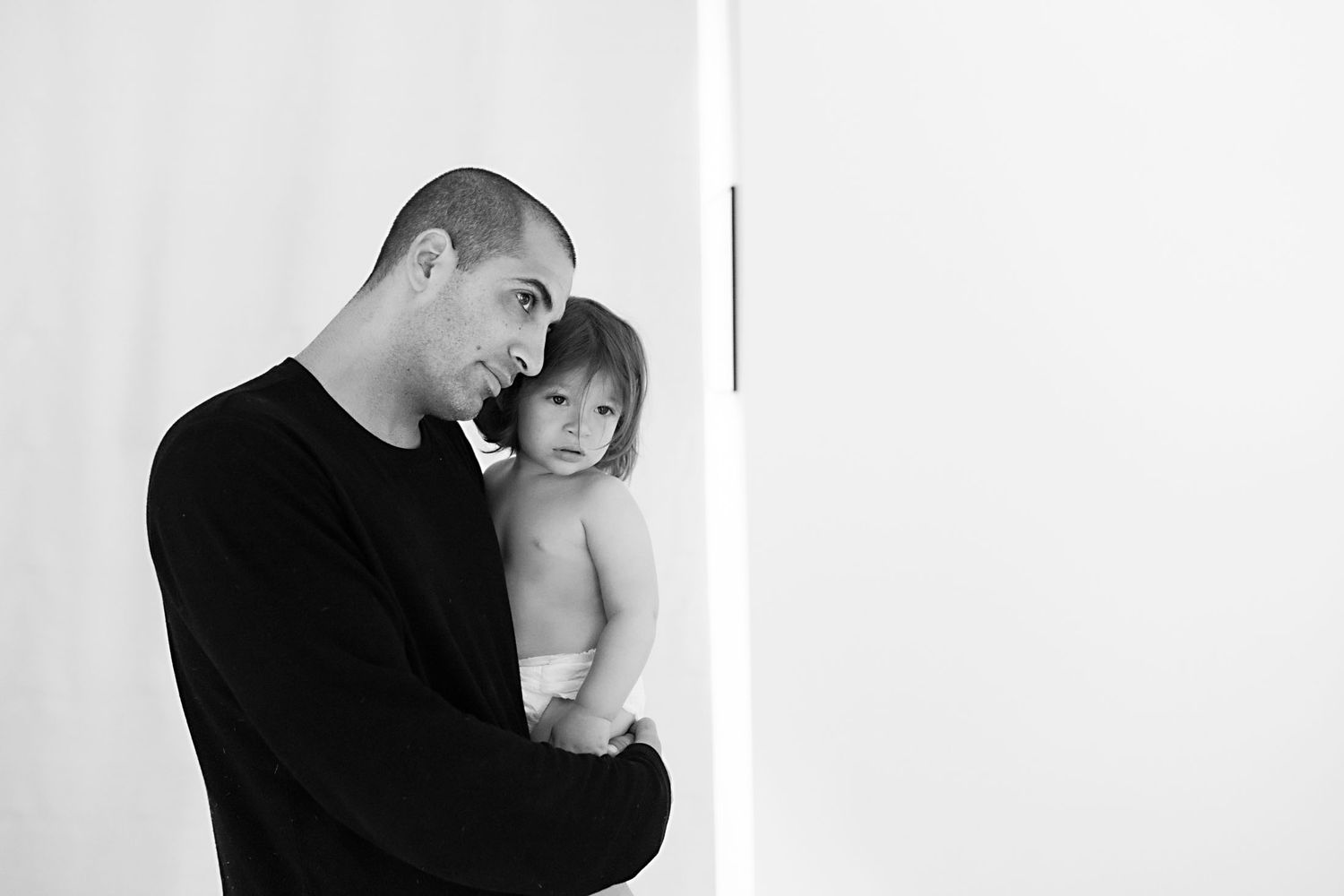 Black and white photo of a man in a black sweater tenderly holding a toddler in a diaper against a white wall.