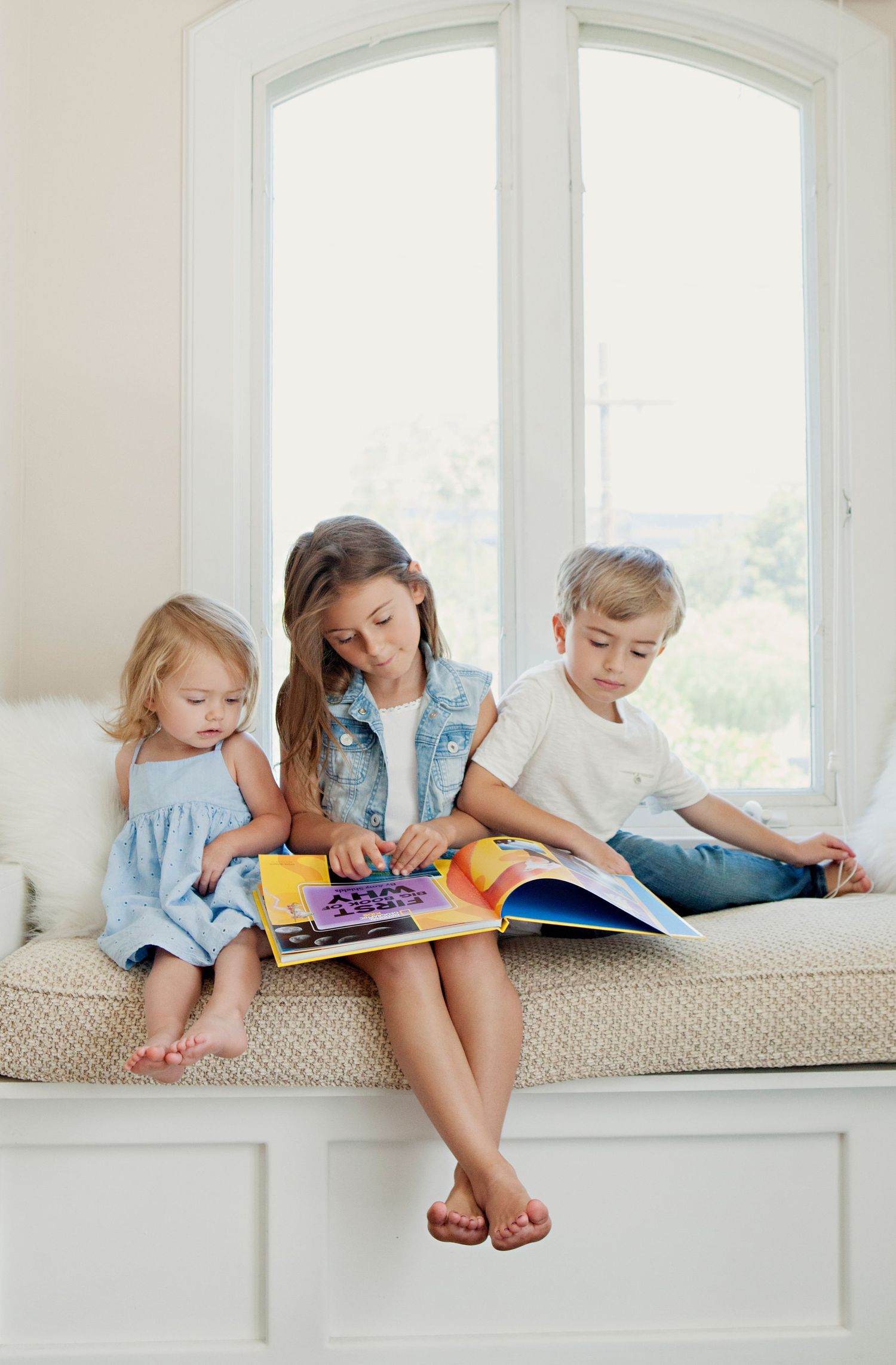 Three young siblings sit on window seat reading a colorful book together in bright natural light.