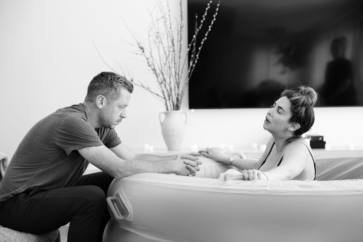 A partner holds hands with a laboring woman in a birthing tub offering comfort during a home water birth.