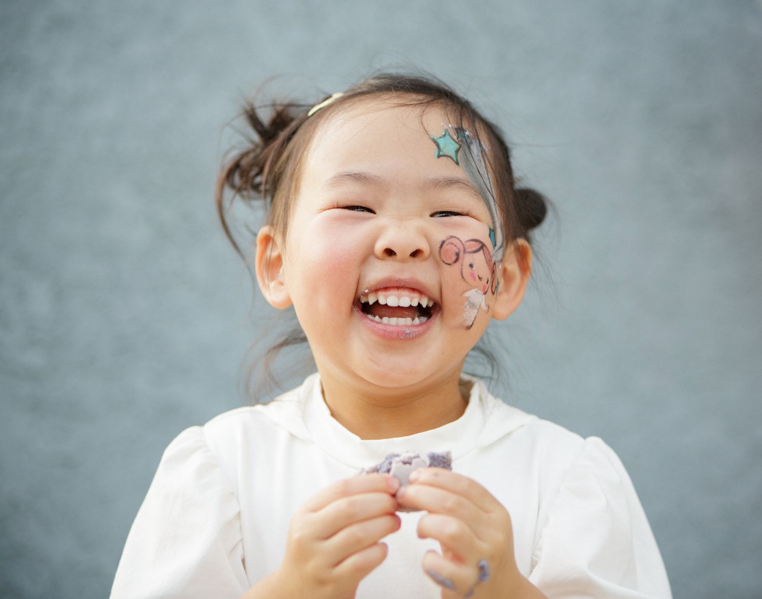 A laughing little girl with star face paint stickers and pigtails wearing a white top.
