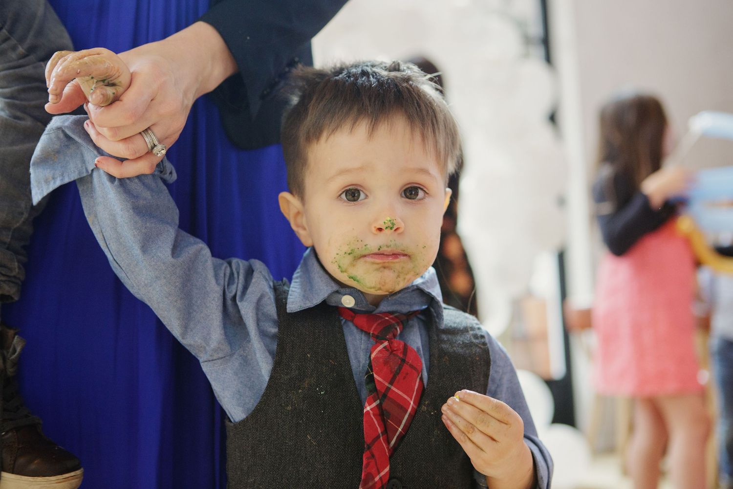 Toddler boy in suit and red tie with food on face held by parent at celebration event.
