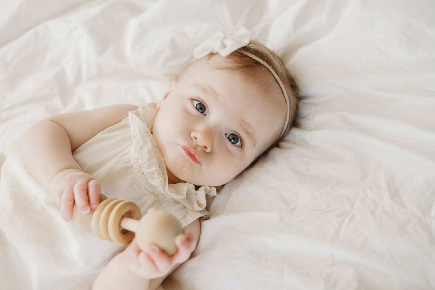 Baby girl in white dress with bow headband lying on white bedding holding a wooden rattle toy.