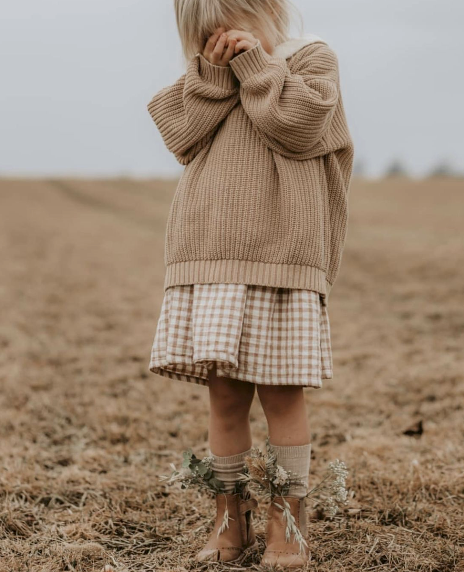 Child in tan knit sweater and gingham skirt with brown boots holds wildflowers in an open field.